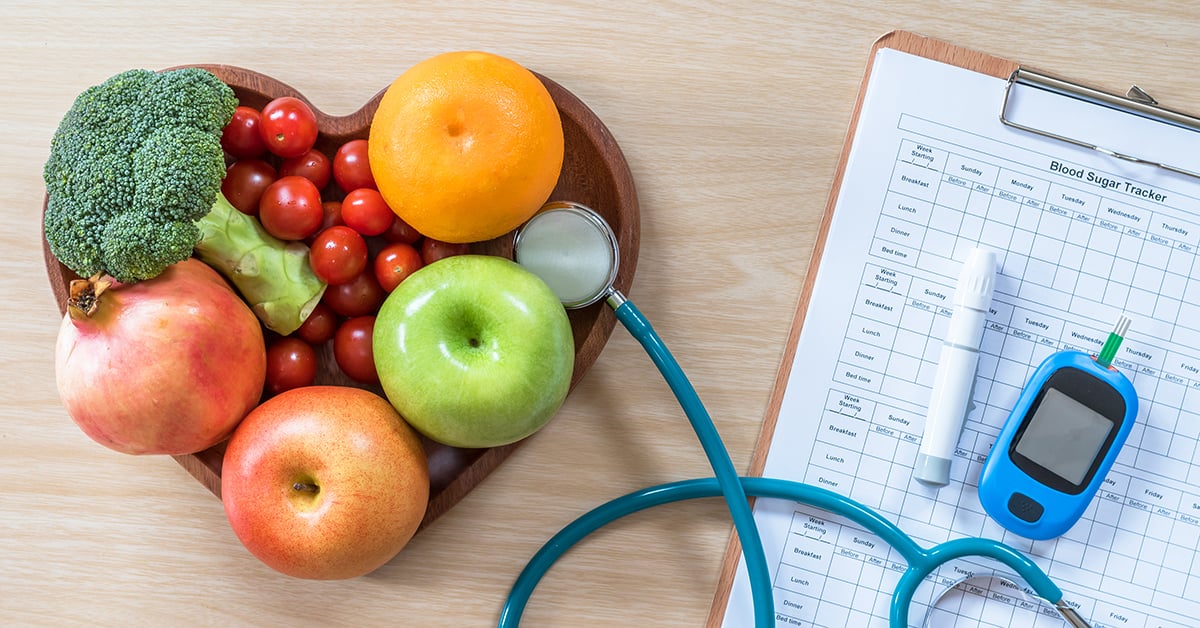 Heart-shaped bowl of fruits and vegetables next to a blood sugar monitor and tracker sheet